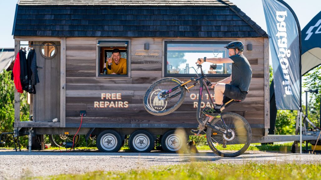 Ein Mountainbiker macht einen Wheelie vor dem Patagonia „Worn Wear" Van, während jemand aus dem Fenster schaut.